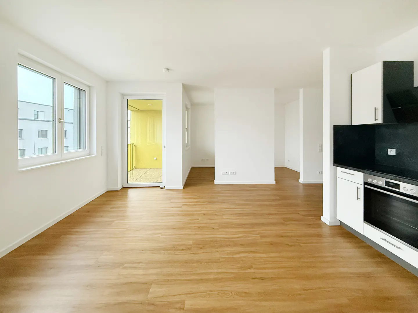 Bright, empty apartment with wood floors, white walls, and a black and white kitchen. A balcony is visible through a sliding glass door.
