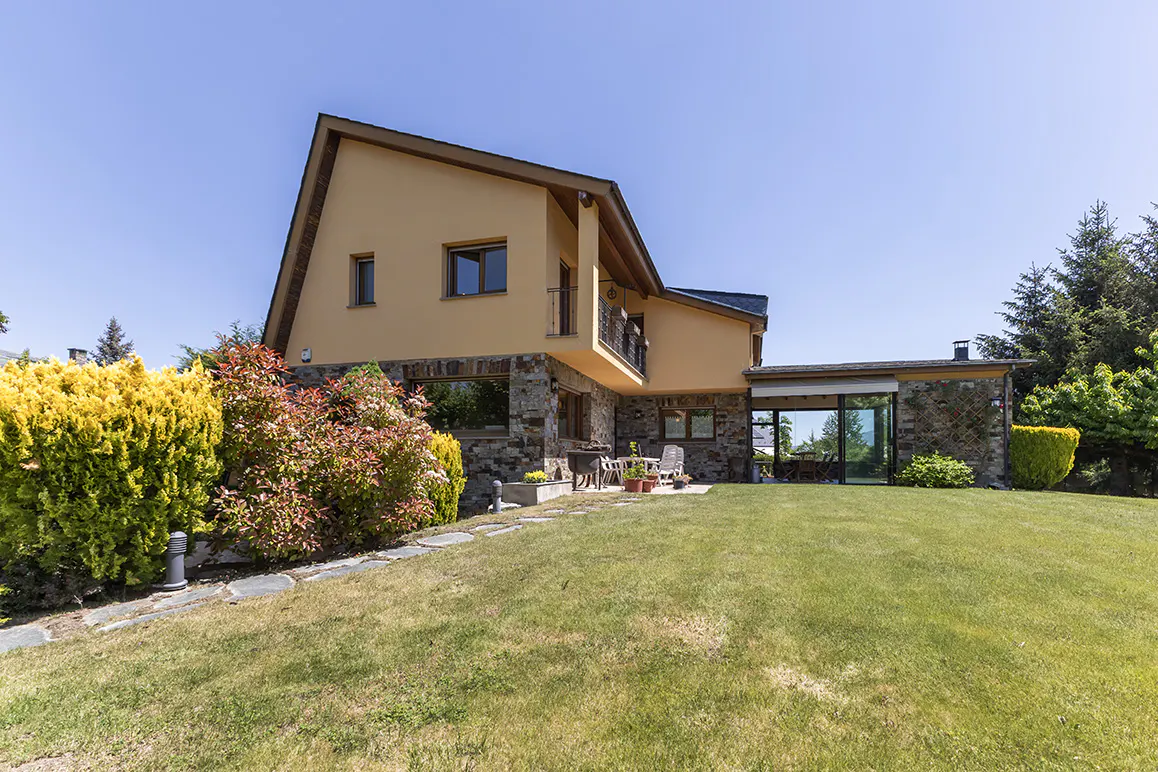 Two-story house with tan stucco and stone exterior, featuring a patio, lush green lawn, and vibrant landscaping.