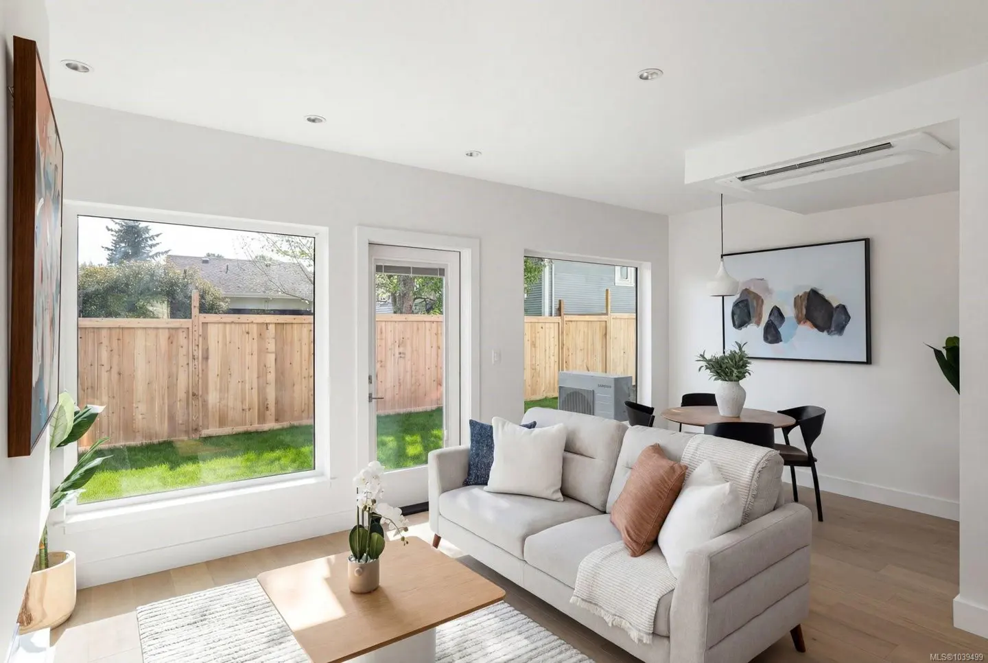 Bright living room with a light gray sofa, wooden coffee table, and dining area overlooking a fenced backyard.