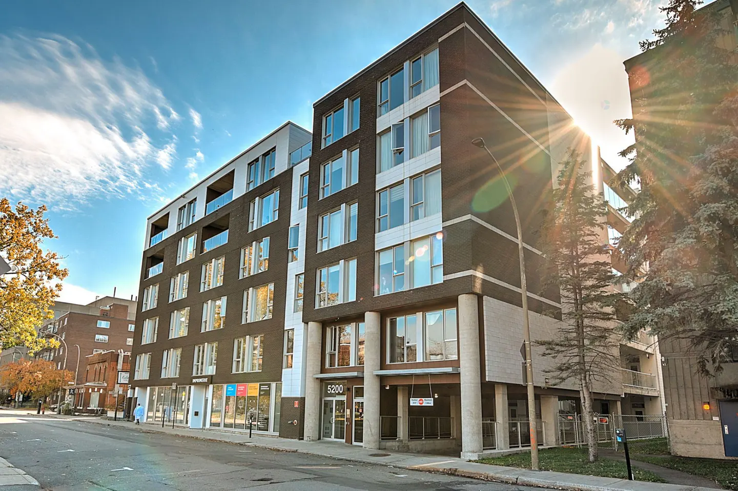 Modern multi-story apartment building with dark brick and white accents, street-level parking, and sunlit blue sky.
