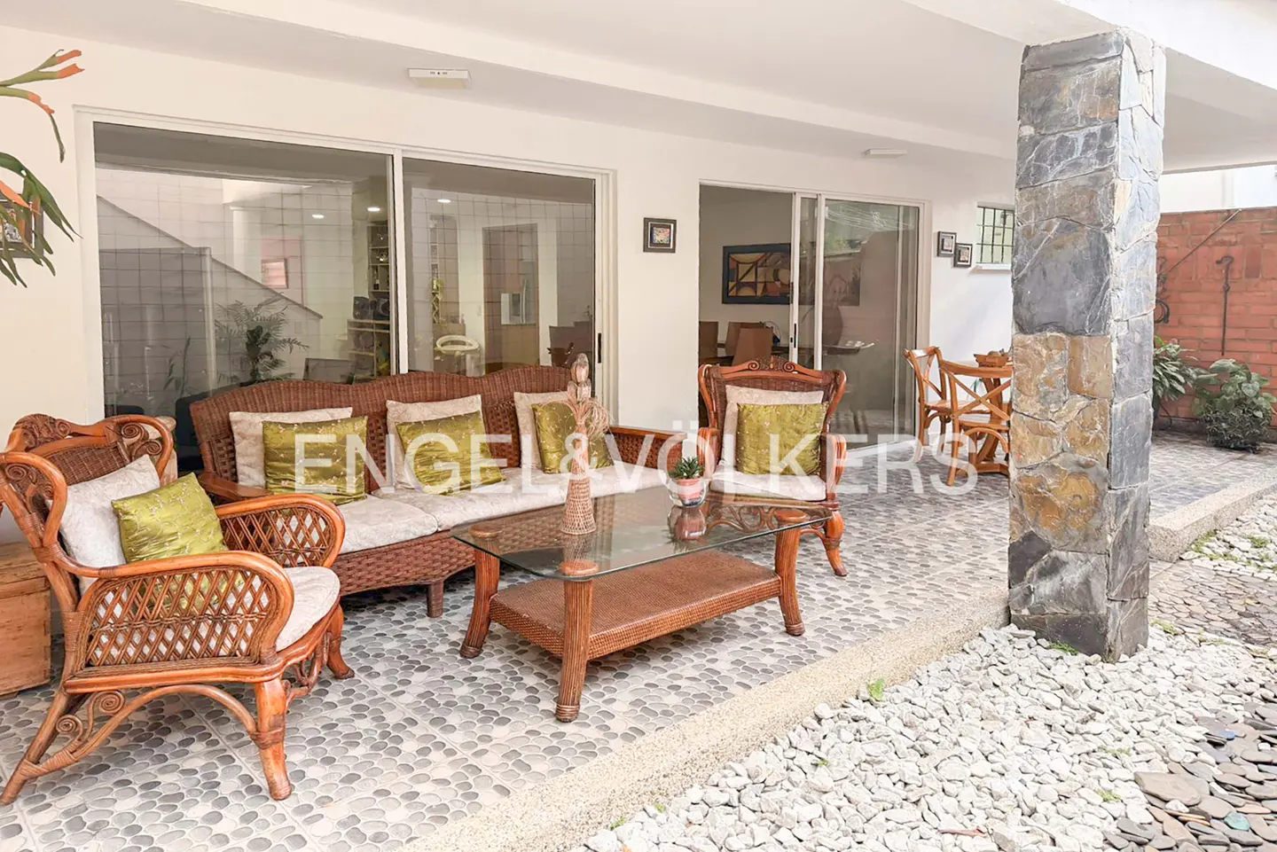 Outdoor patio with wicker furniture, including a sofa, chairs, and a glass-topped coffee table. The floor is tiled with small stones. Sliding glass doors lead inside.