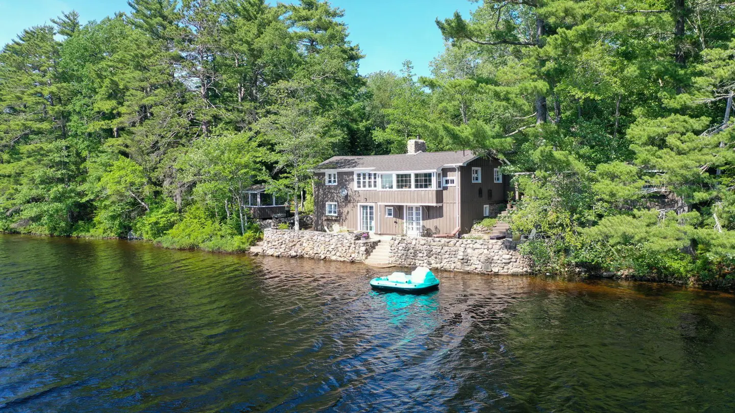Lakefront home with a stone foundation and a teal inflatable boat floating in the water. Trees surround the property.