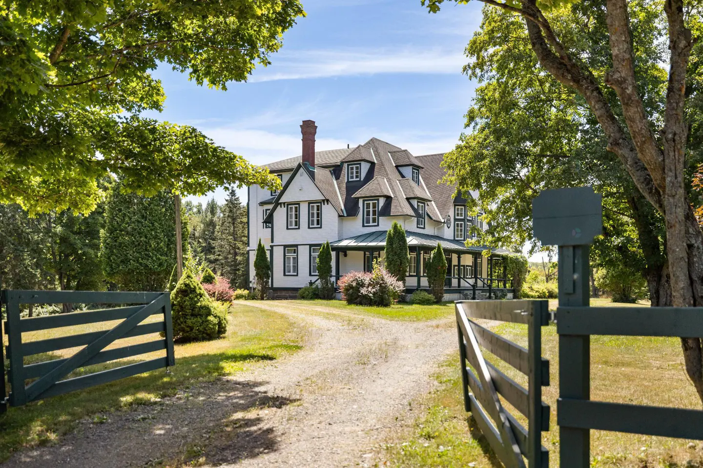 Exterior view of a large white house with black trim, seen through an open green wooden gate. A gravel driveway leads to the house, surrounded by trees.
