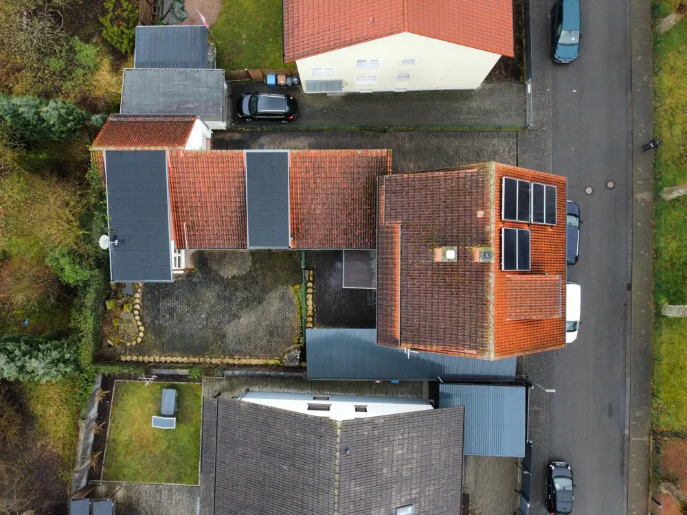 Aerial view of a house with red tile roof and solar panels, surrounded by trees and other houses.