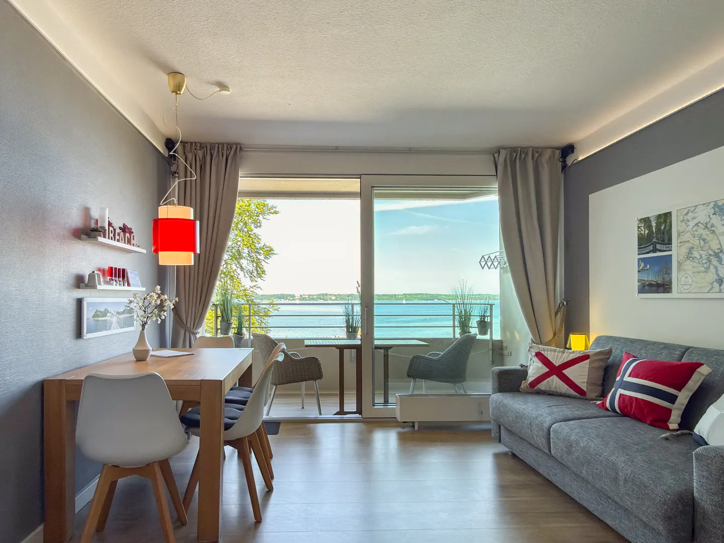 Modern living room with a dining area, grey sofa, and large sliding glass doors opening to a balcony with a sea view.