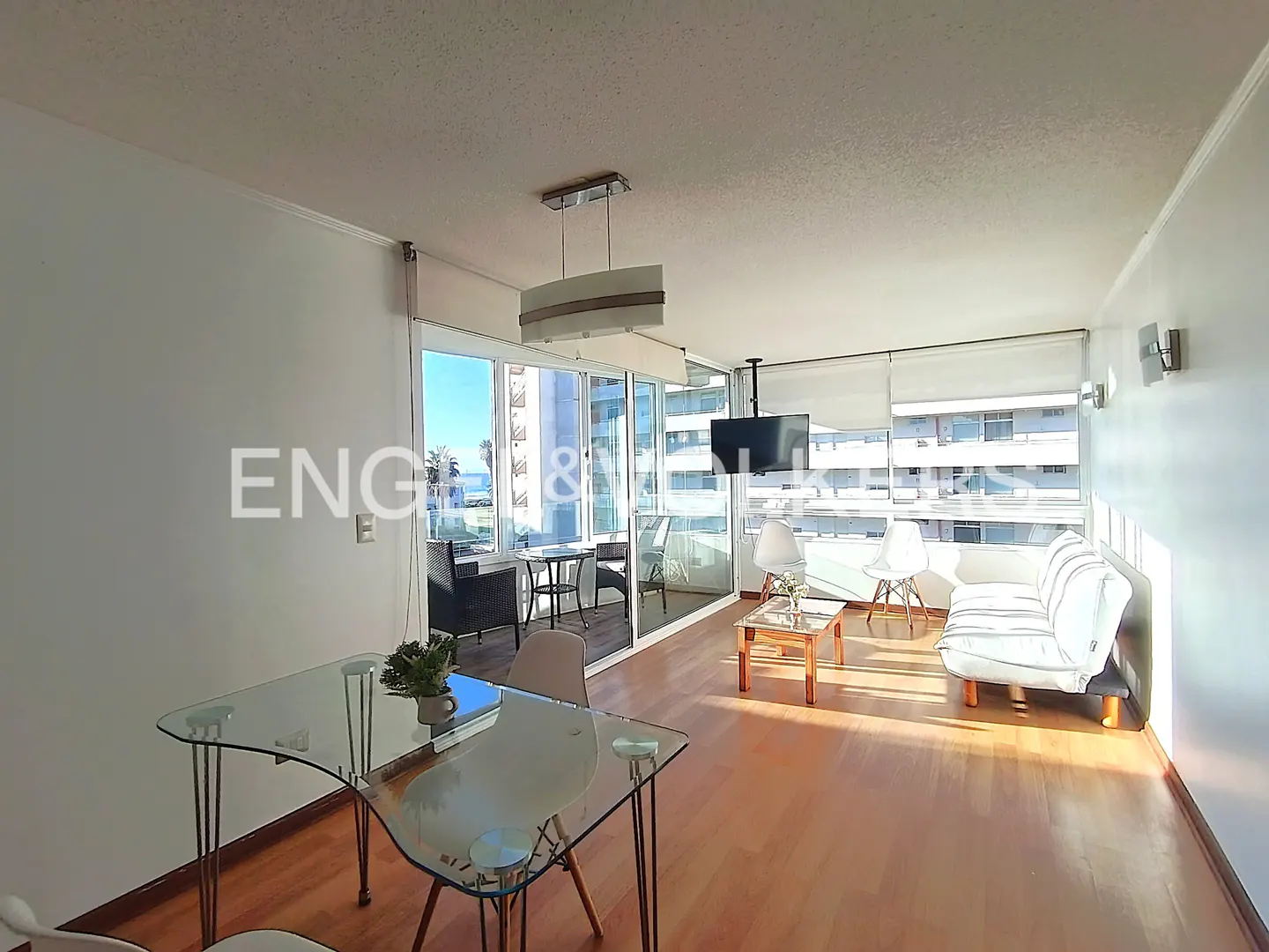 Bright living room with wood floors, glass table, white sofa, and balcony with ocean view.
