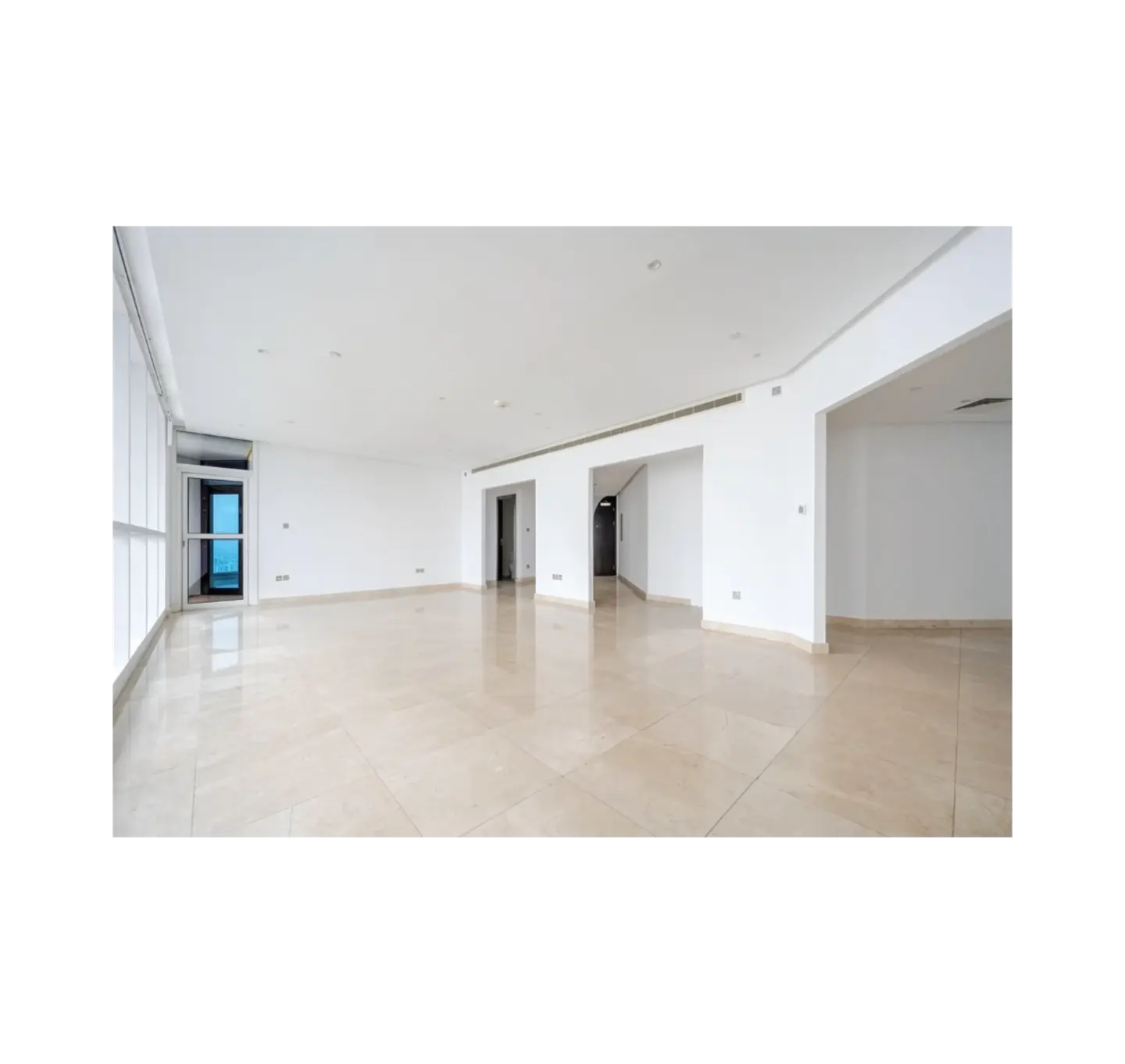 An empty, bright room with white walls and beige tile floors. There are windows on the left and doorways on the right.