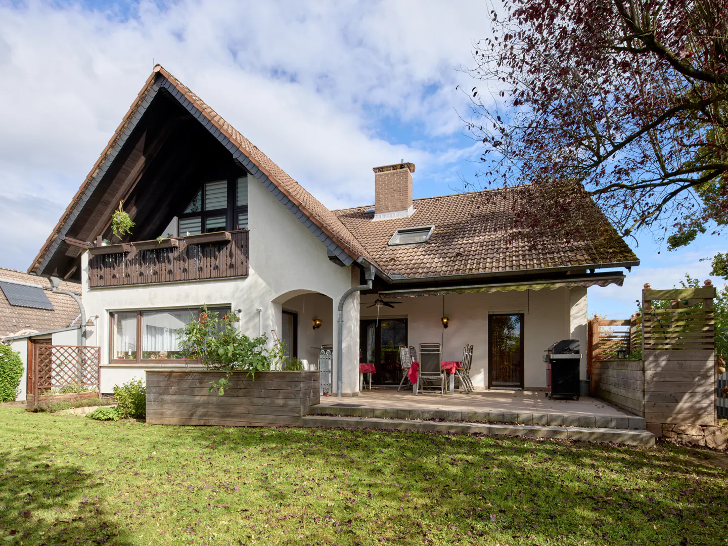 Two-story white house with a brown roof, balcony, and patio with chairs and a grill, surrounded by a green lawn and trees.