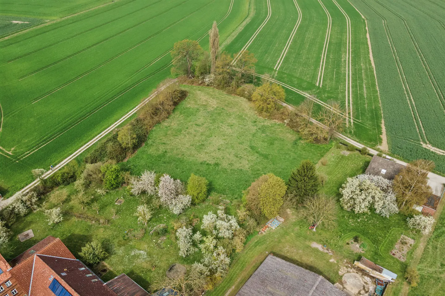 Aerial view of a green field surrounded by trees and farmland, with a building visible in the lower left corner.