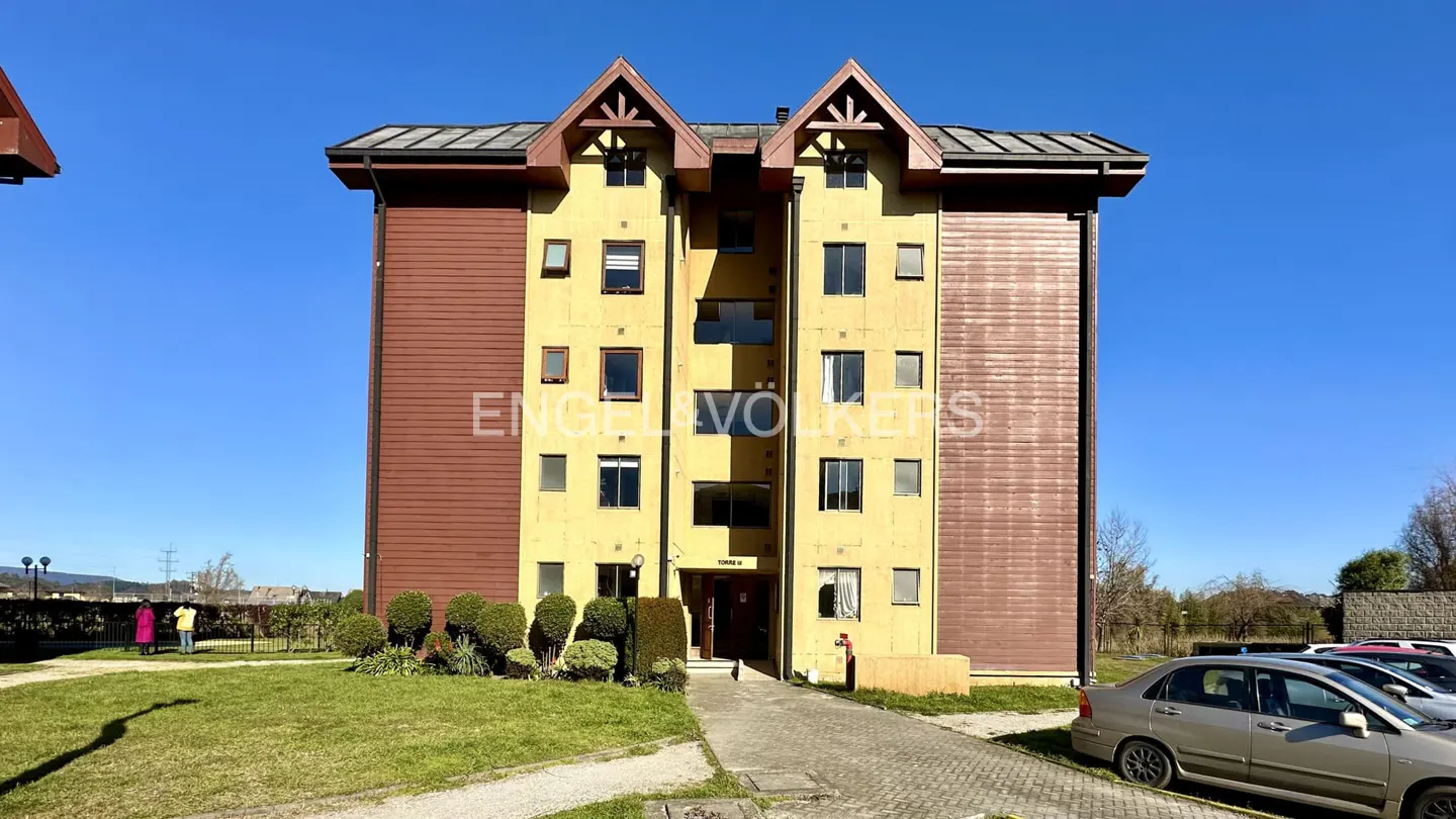 Exterior view of a yellow and brown apartment building with a green lawn and parked cars under a clear blue sky.