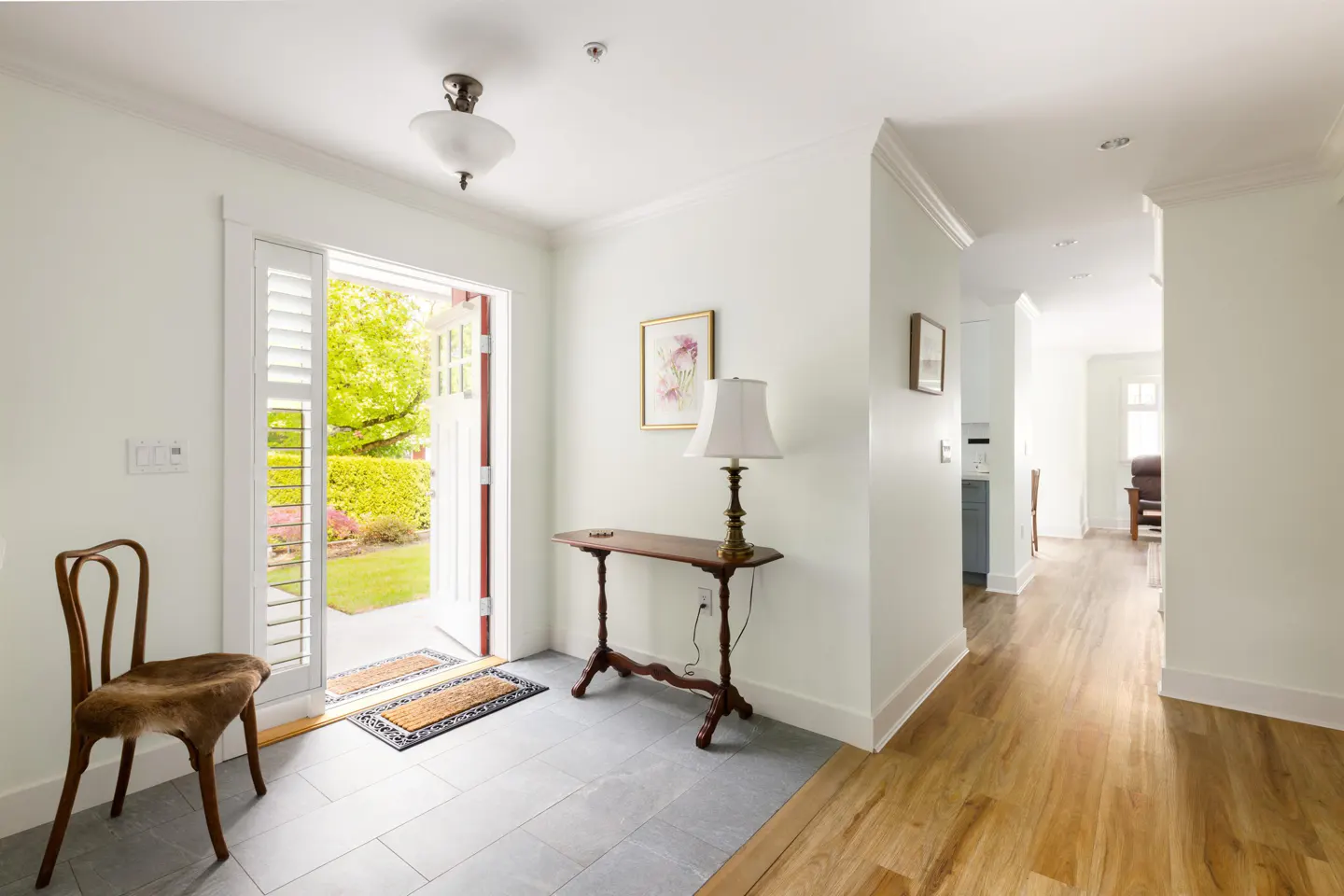 Bright foyer with white walls, gray tile, and wood floors. An open door leads to a green garden. A wooden chair and table furnish the space.