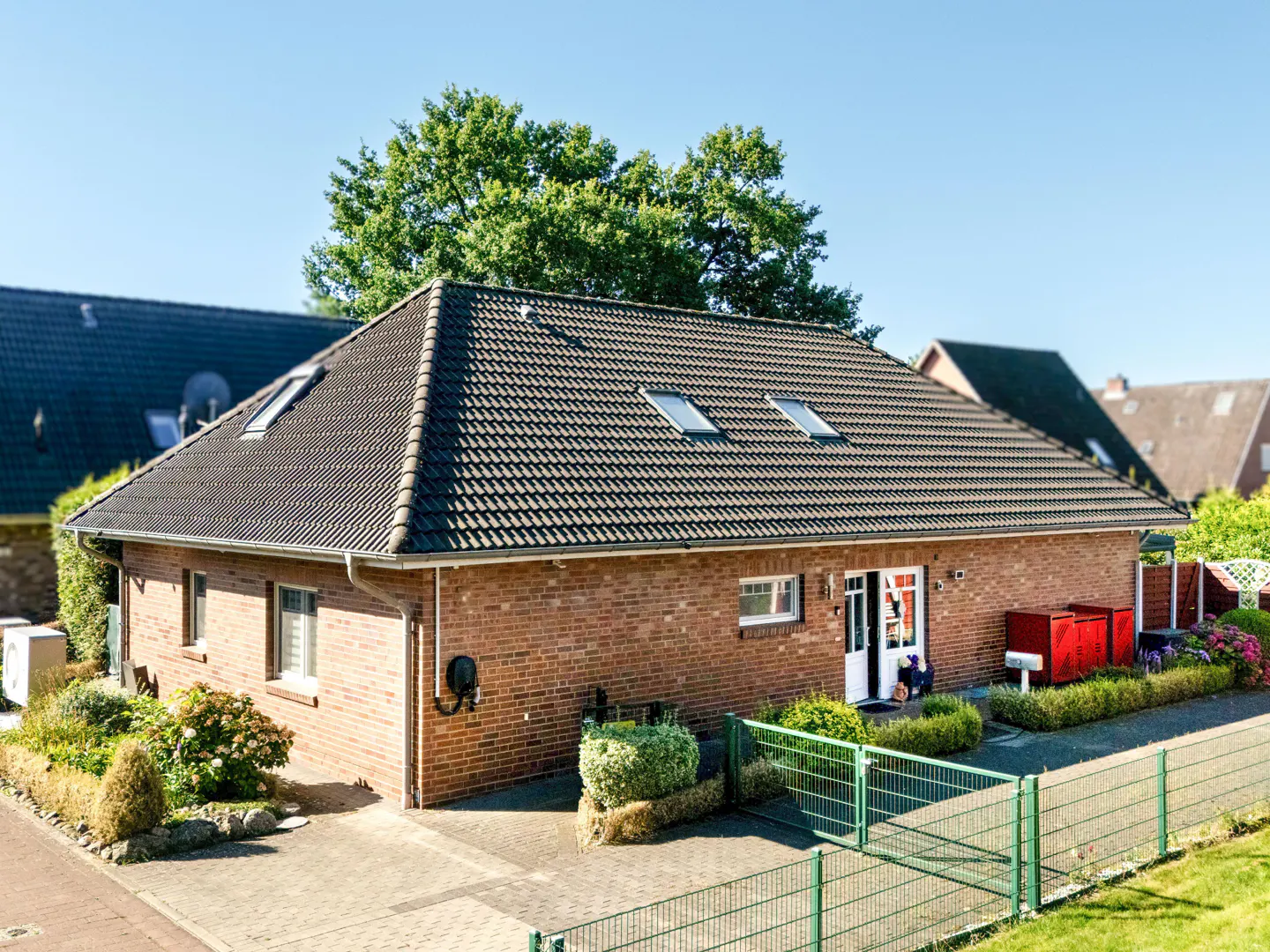 Single-story red brick house with a dark tiled roof, skylights, and a paved driveway behind a green metal fence.