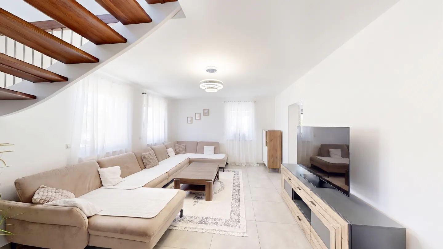 Modern living room with a beige sectional sofa, wooden coffee table, TV console, and open wooden staircase.