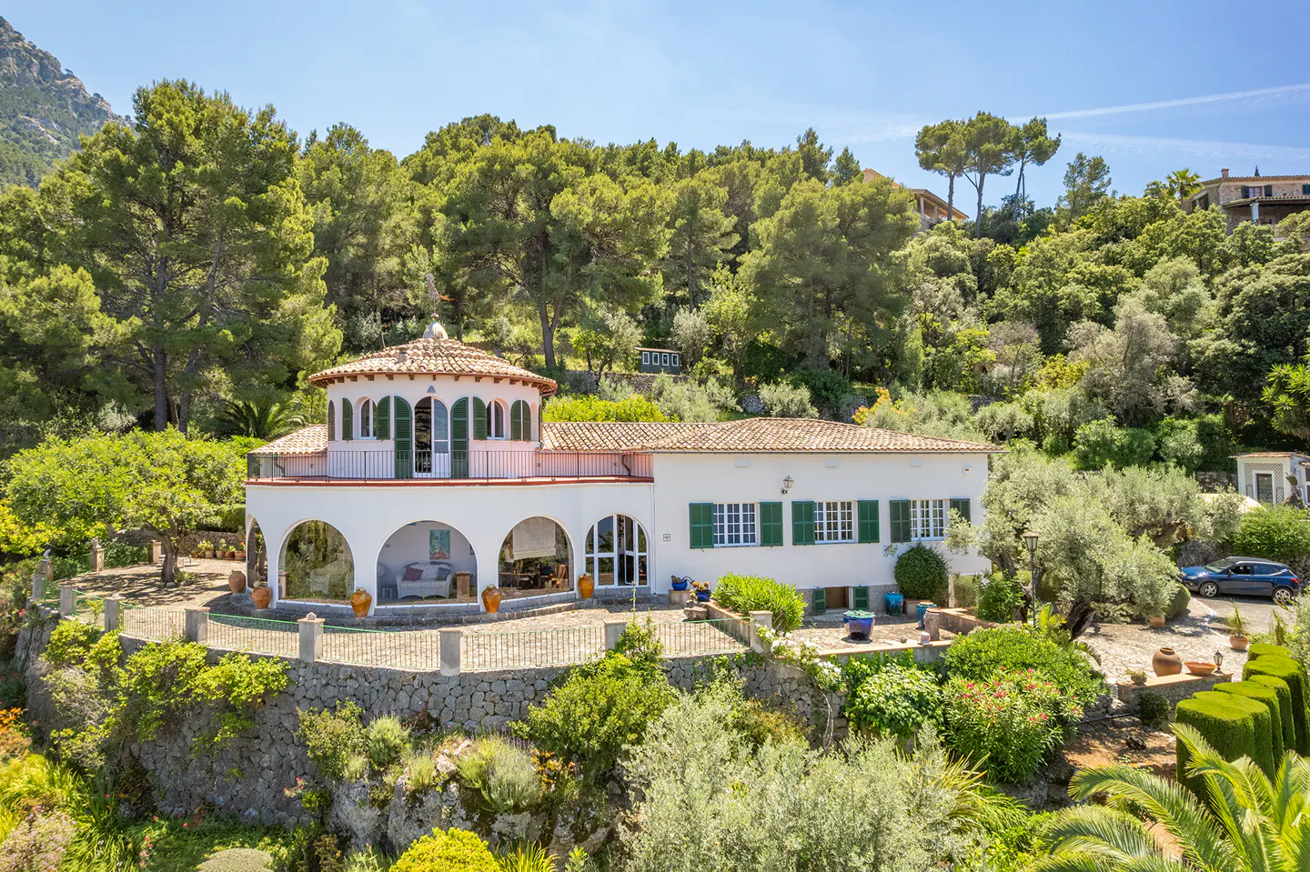 White Mediterranean villa with a circular terrace, green shutters, and tiled roof, nestled in a lush, hilly landscape.