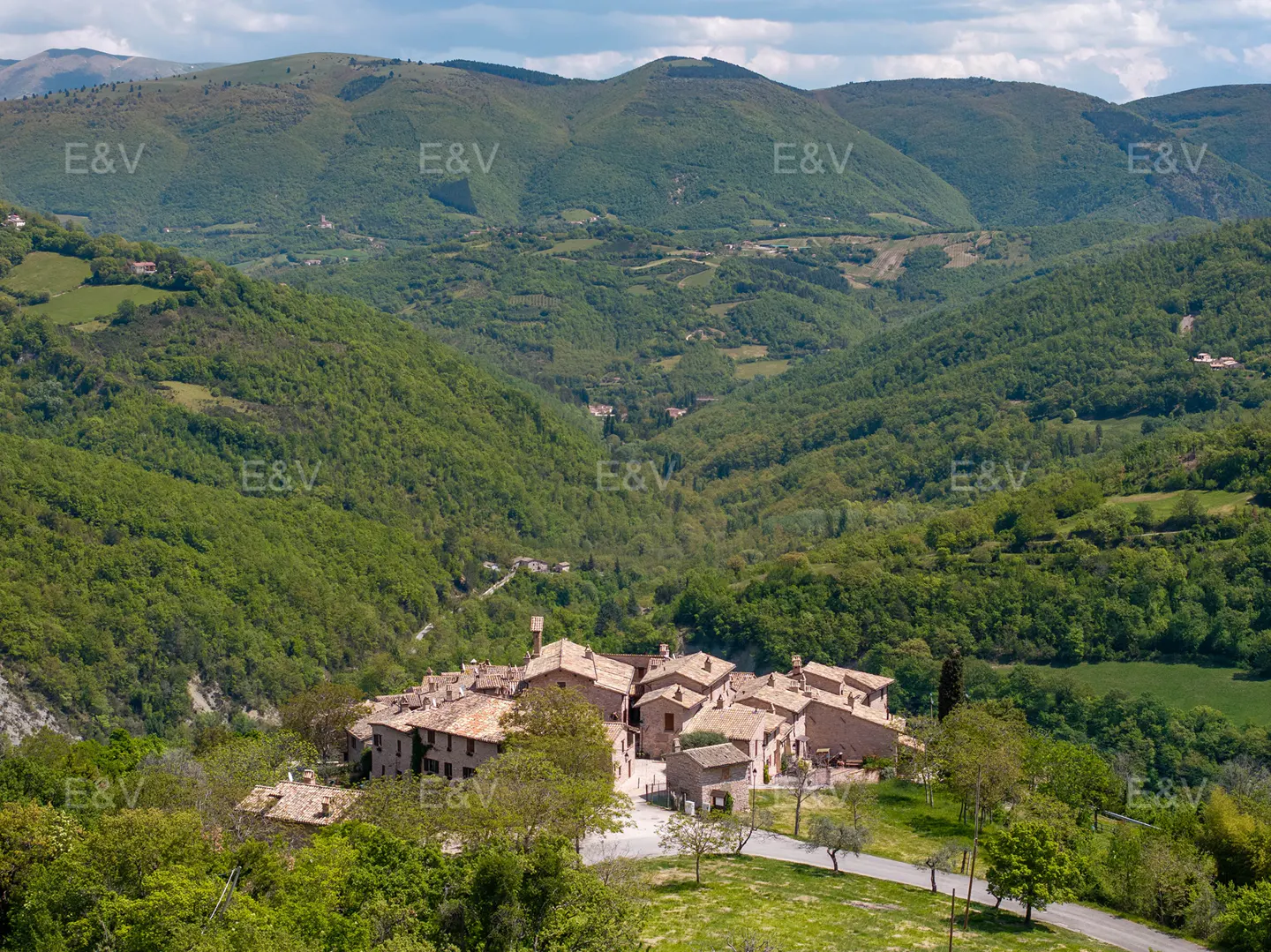 A hilltop village with red-tiled roofs nestled in a green valley, surrounded by forested hills under a partly cloudy sky.