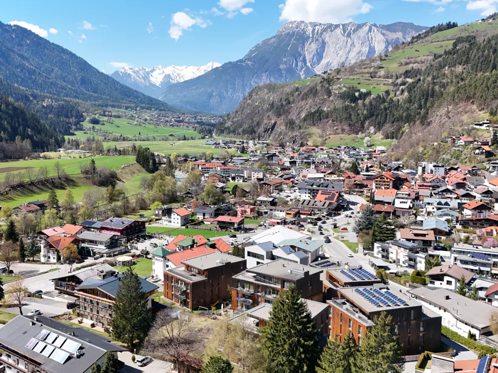 Scenic view of a European town nestled in a valley, surrounded by green hills and snow-capped mountains under a blue sky.