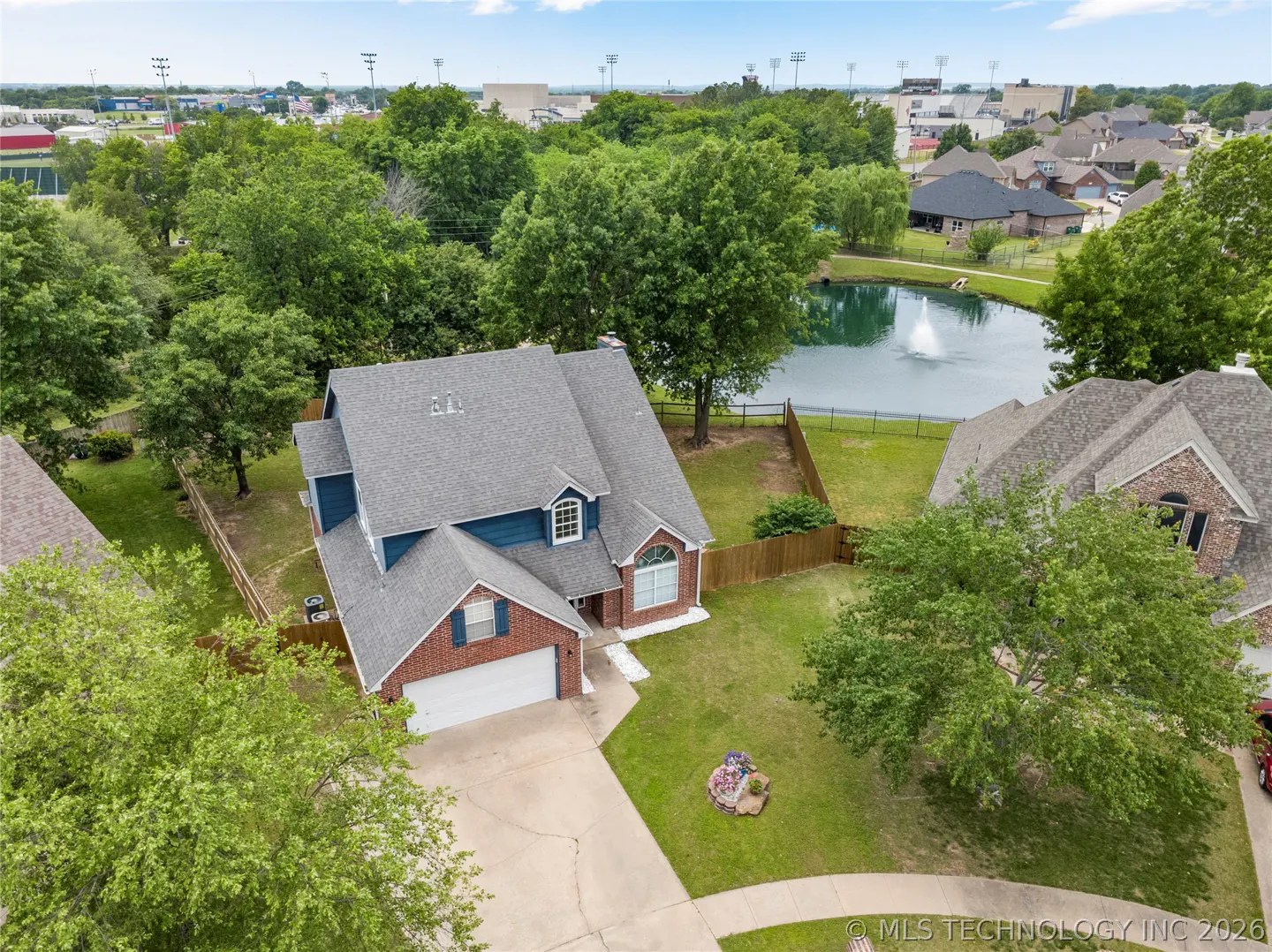Aerial view of a two-story house with blue siding, brick accents, and a gray roof, surrounded by green trees and a pond.