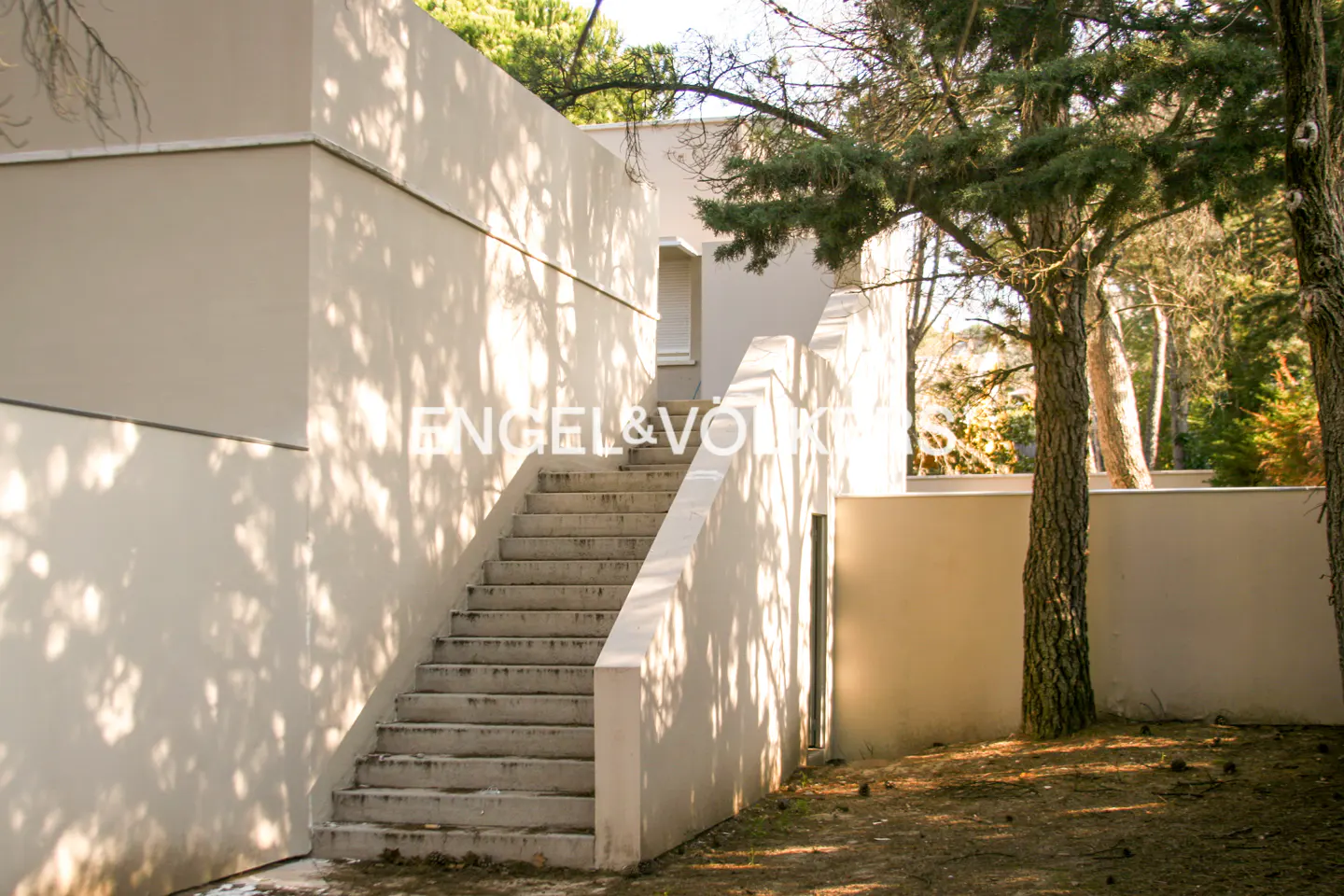 Exterior view of a modern beige house with an outdoor staircase, surrounded by trees and dappled sunlight.