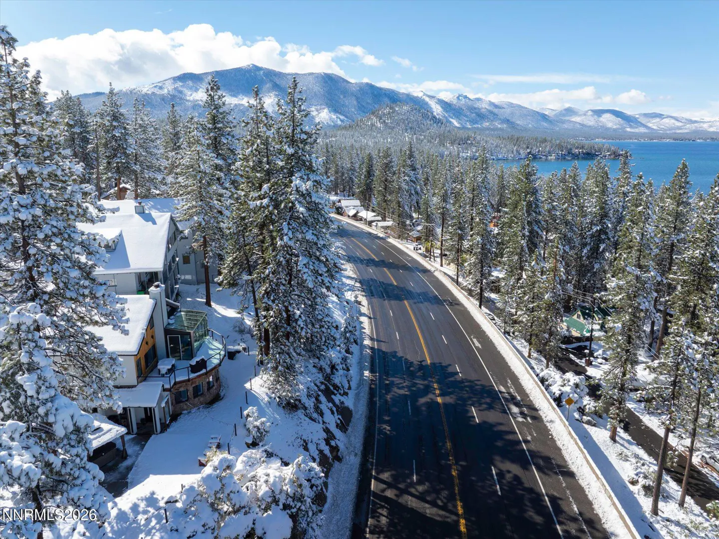 Aerial view of a snow-covered road lined with pine trees, houses, and a lake with mountains in the background.