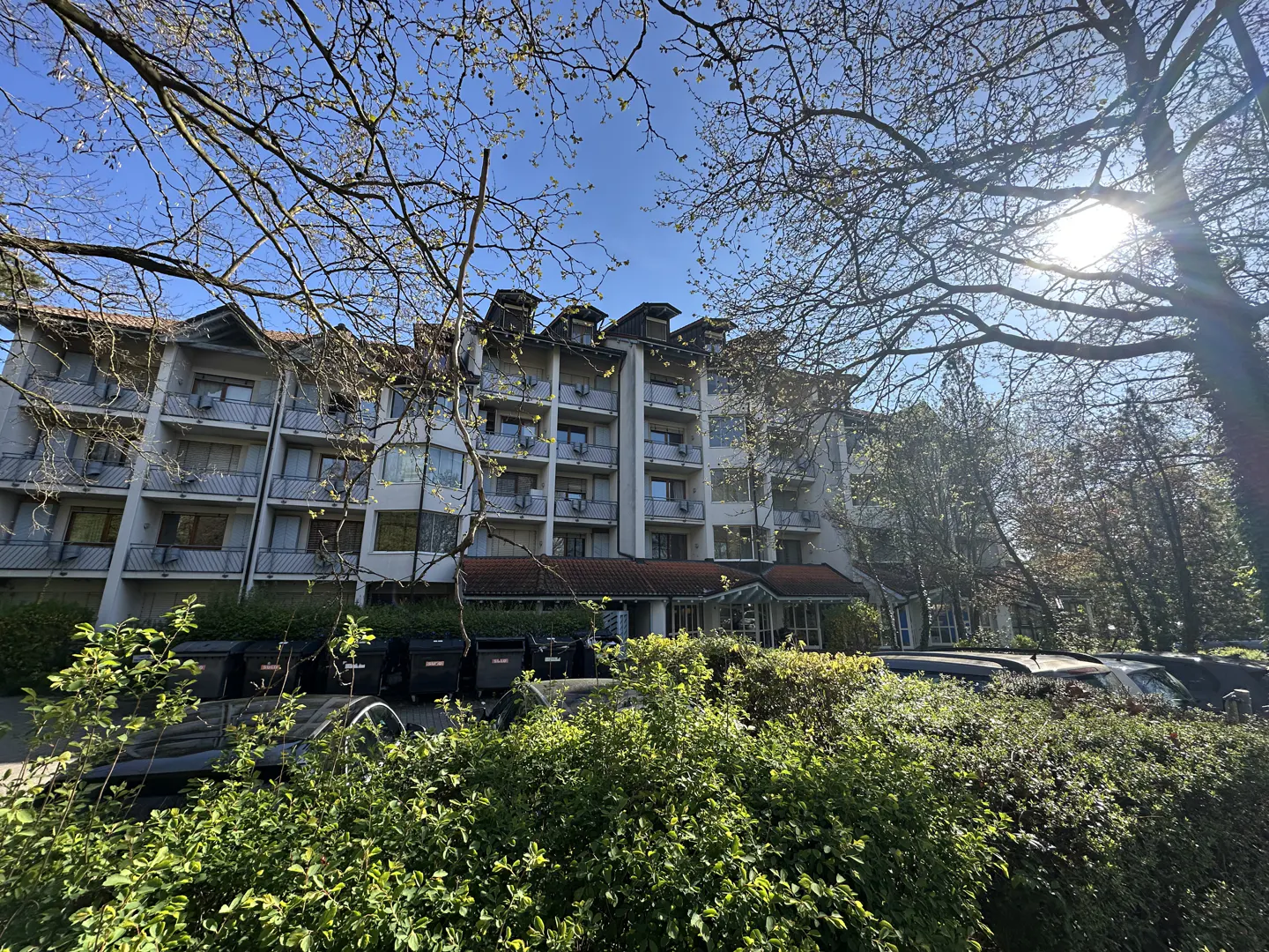 Exterior view of a multi-story building with balconies, surrounded by trees and greenery on a sunny day.
