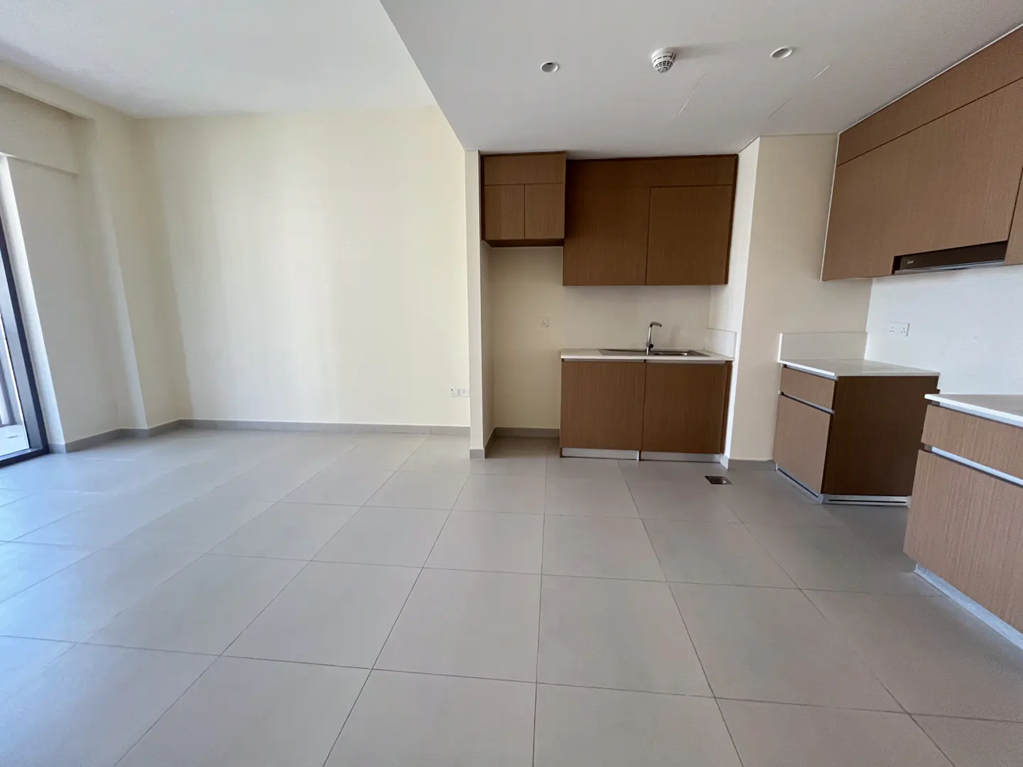 Modern open-plan kitchen with brown cabinets, white countertops, and light grey tiled floors in a bright, empty apartment.