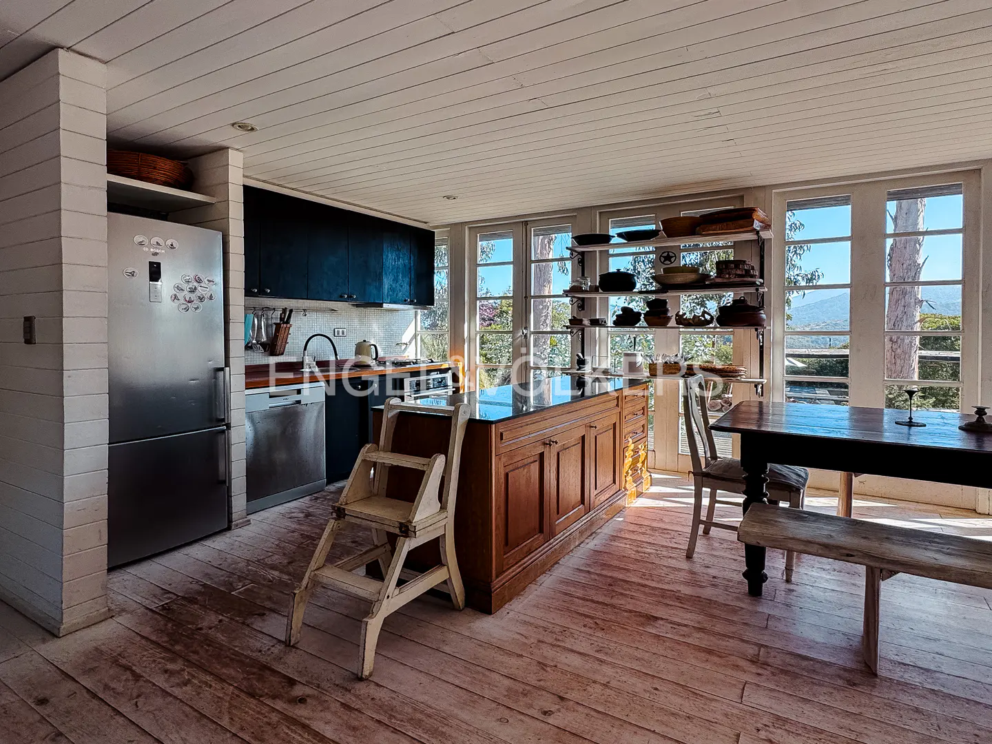 A bright kitchen with a wood island, black countertops, and a white step stool. A stainless steel fridge and dishwasher are visible. Large windows offer a view of trees.