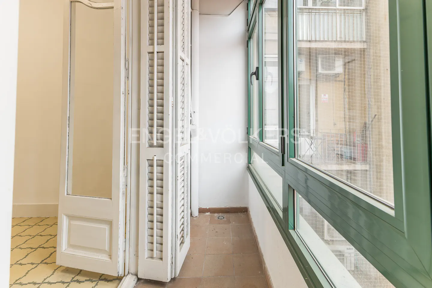 A bright sunroom with white shutters and green-framed windows. Outside, a building is visible.