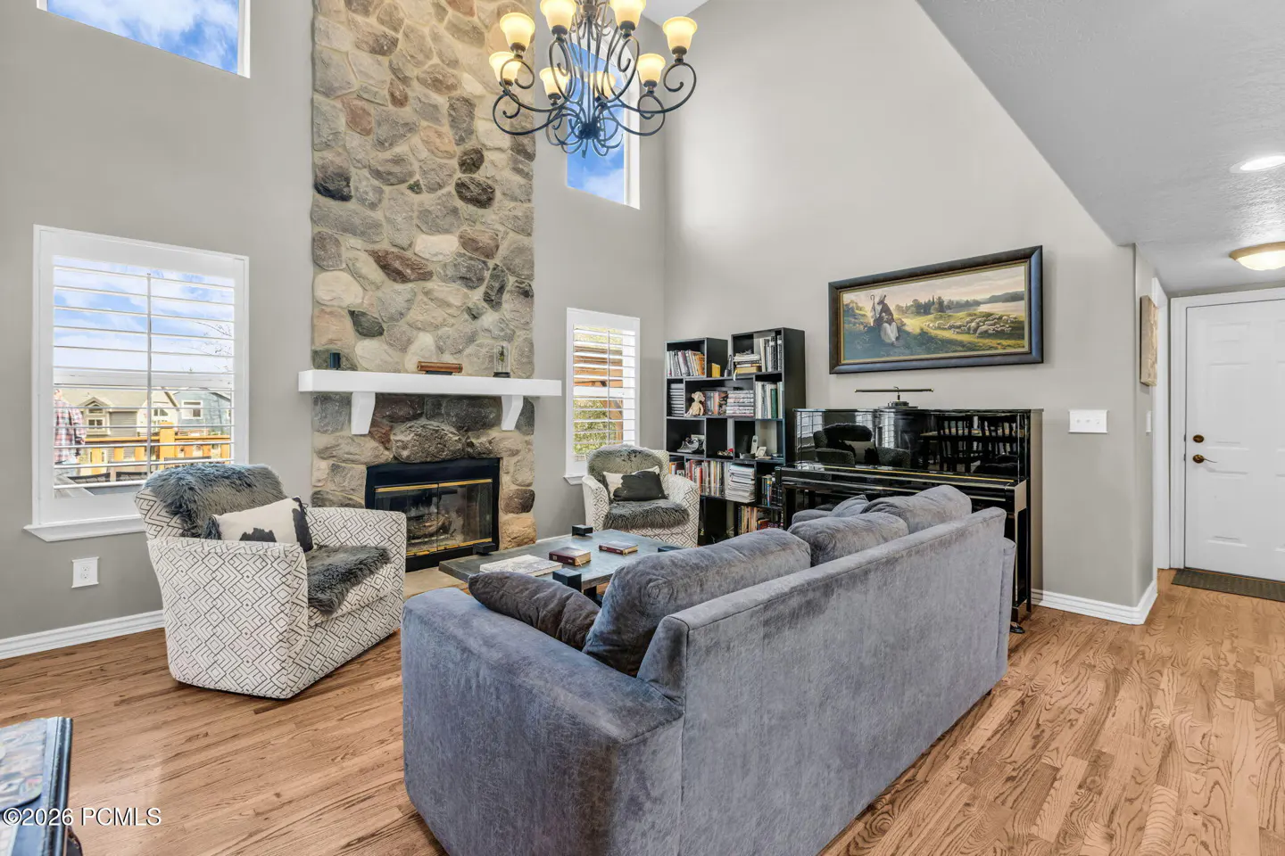 Living room with stone fireplace, gray sofa, piano, and hardwood floors. A chandelier hangs from the high ceiling.