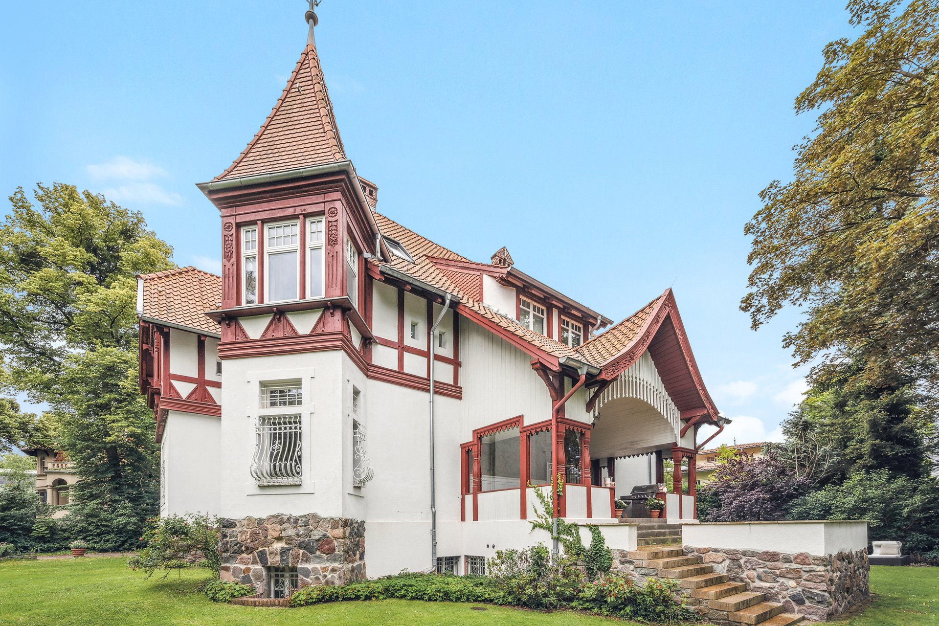 Exterior view of a white, two-story house with red trim, a turret, and a stone foundation, surrounded by green lawn and trees.