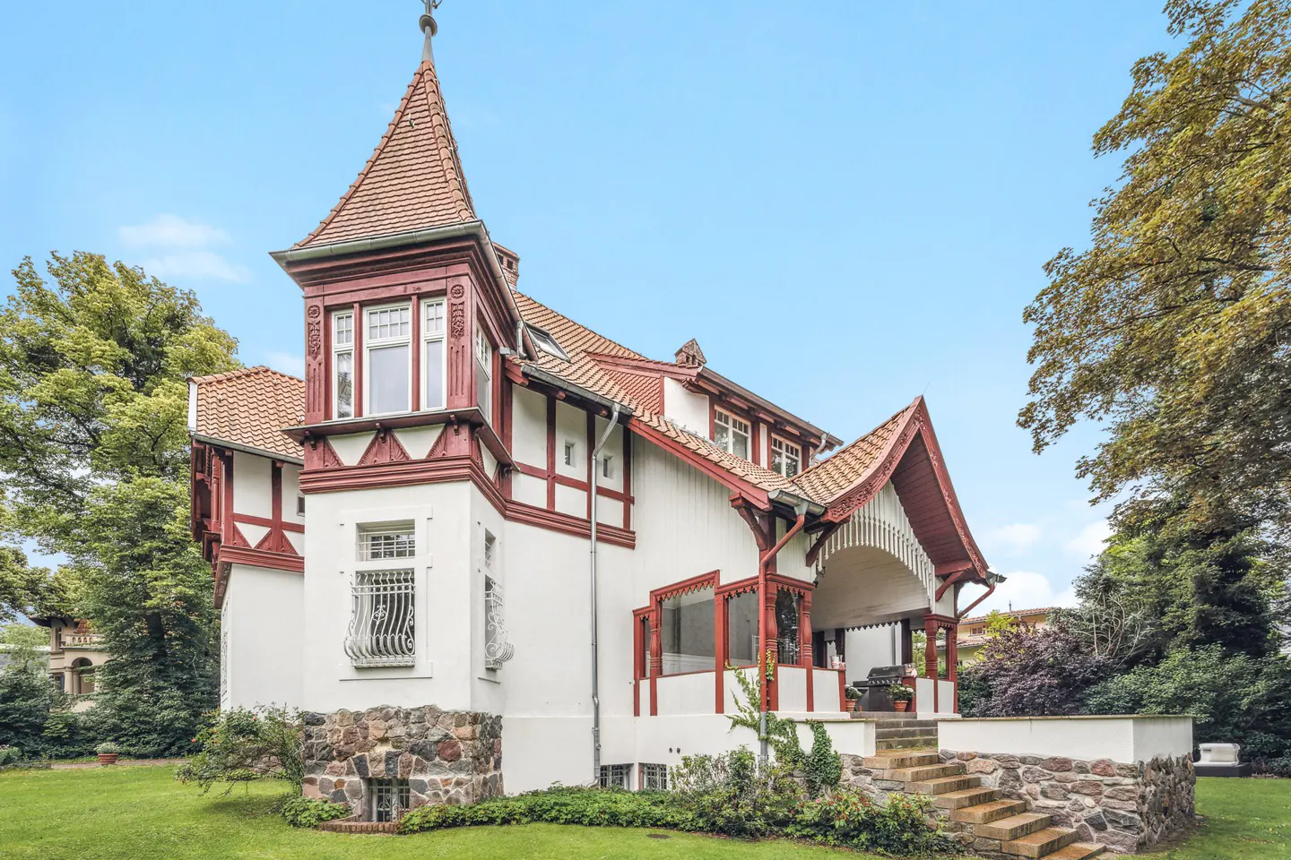 Exterior view of a white, two-story house with red trim, a turret, and a stone foundation, surrounded by green lawn and trees.