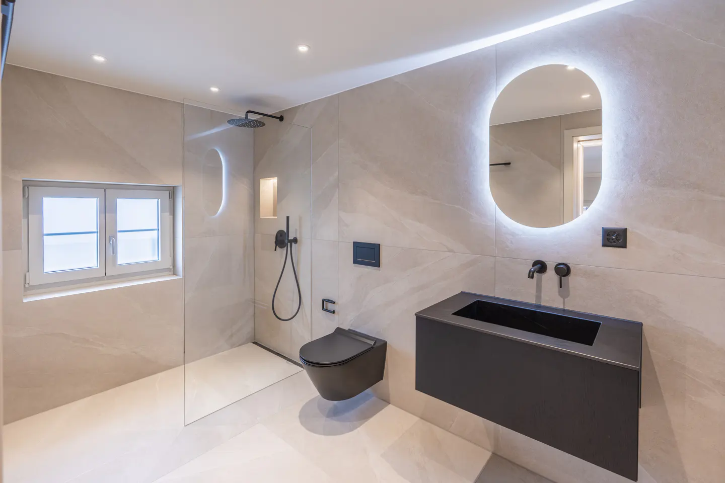 Modern bathroom with beige tile walls, black toilet and sink, and a lighted oval mirror. A window provides natural light.