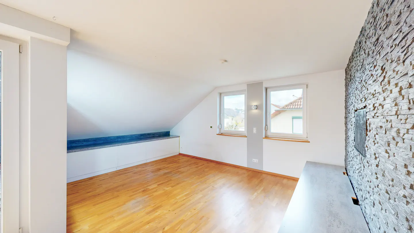 Bright attic room with hardwood floors, white walls, and two windows. A stone accent wall adds texture. Built-in storage under the sloped ceiling.