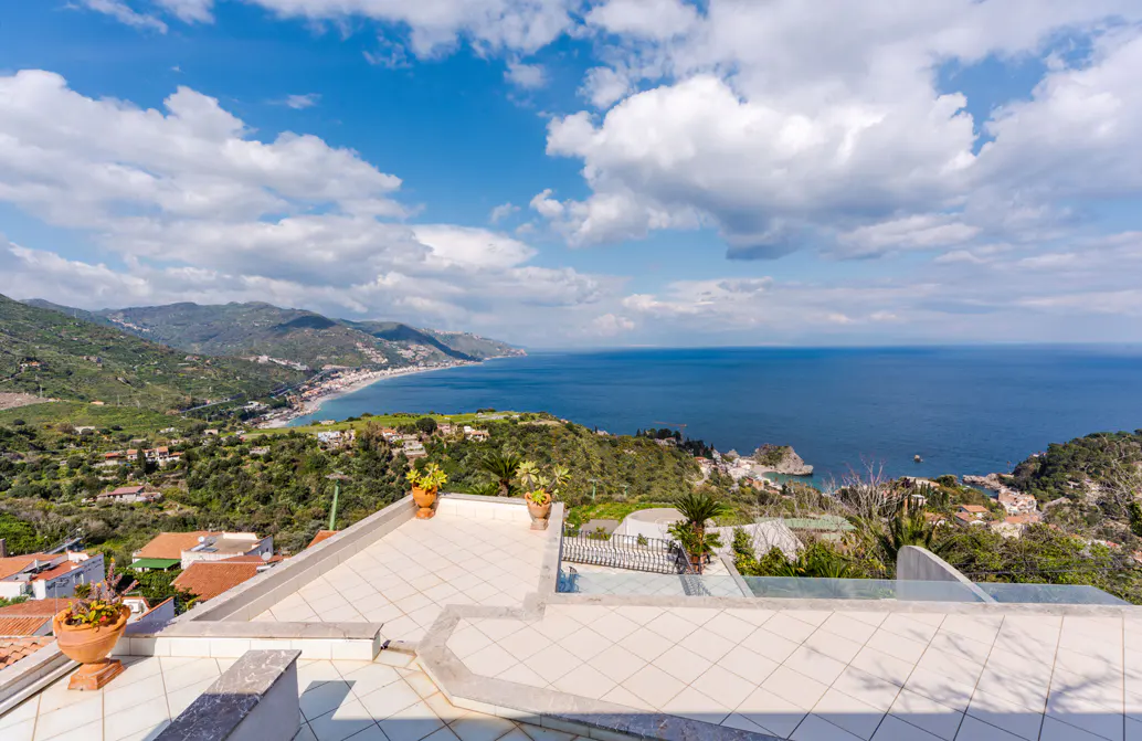 View from a tiled terrace overlooking the Ionian Sea, mountains, and a blue sky with white clouds.