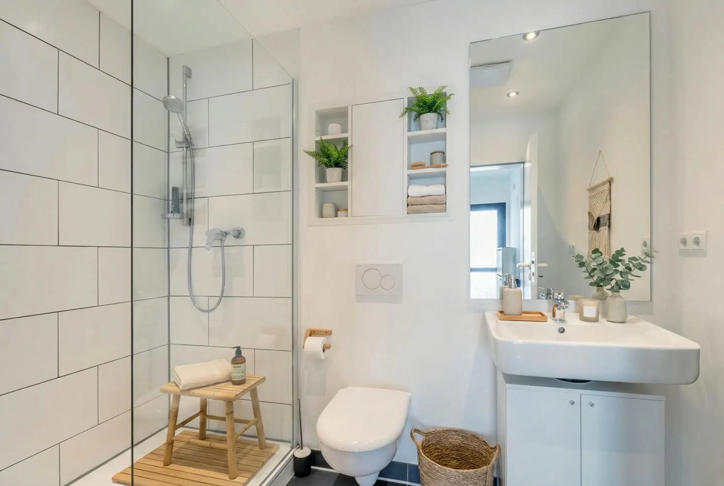 Bright bathroom with white walls, a glass-enclosed shower, toilet, and sink with a mirror. Green plants add a touch of nature.
