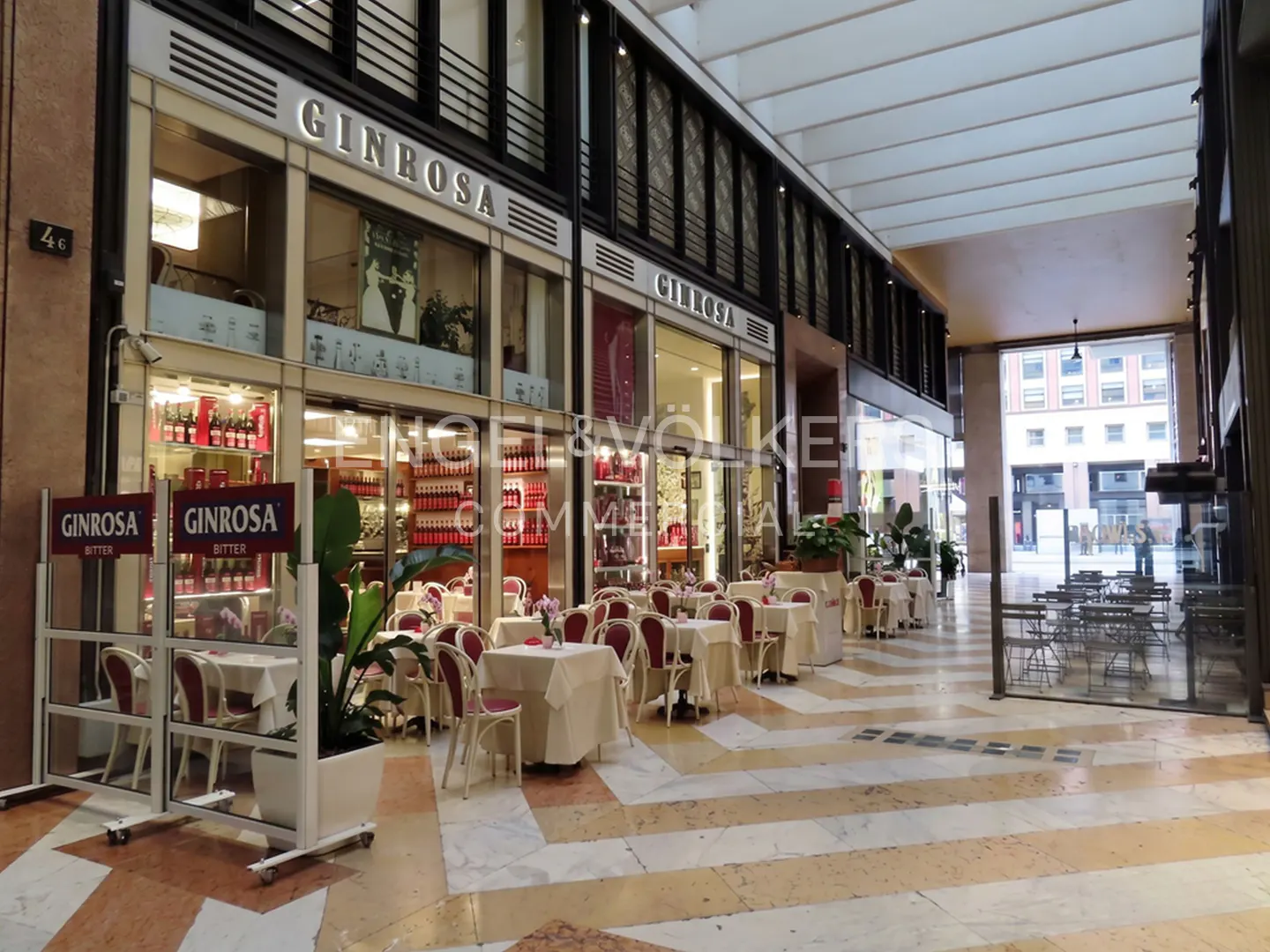 Outdoor seating area of Ginrosa restaurant with tables, chairs, and a marble floor. The restaurant has a glass facade and a sign with the name "Ginrosa".