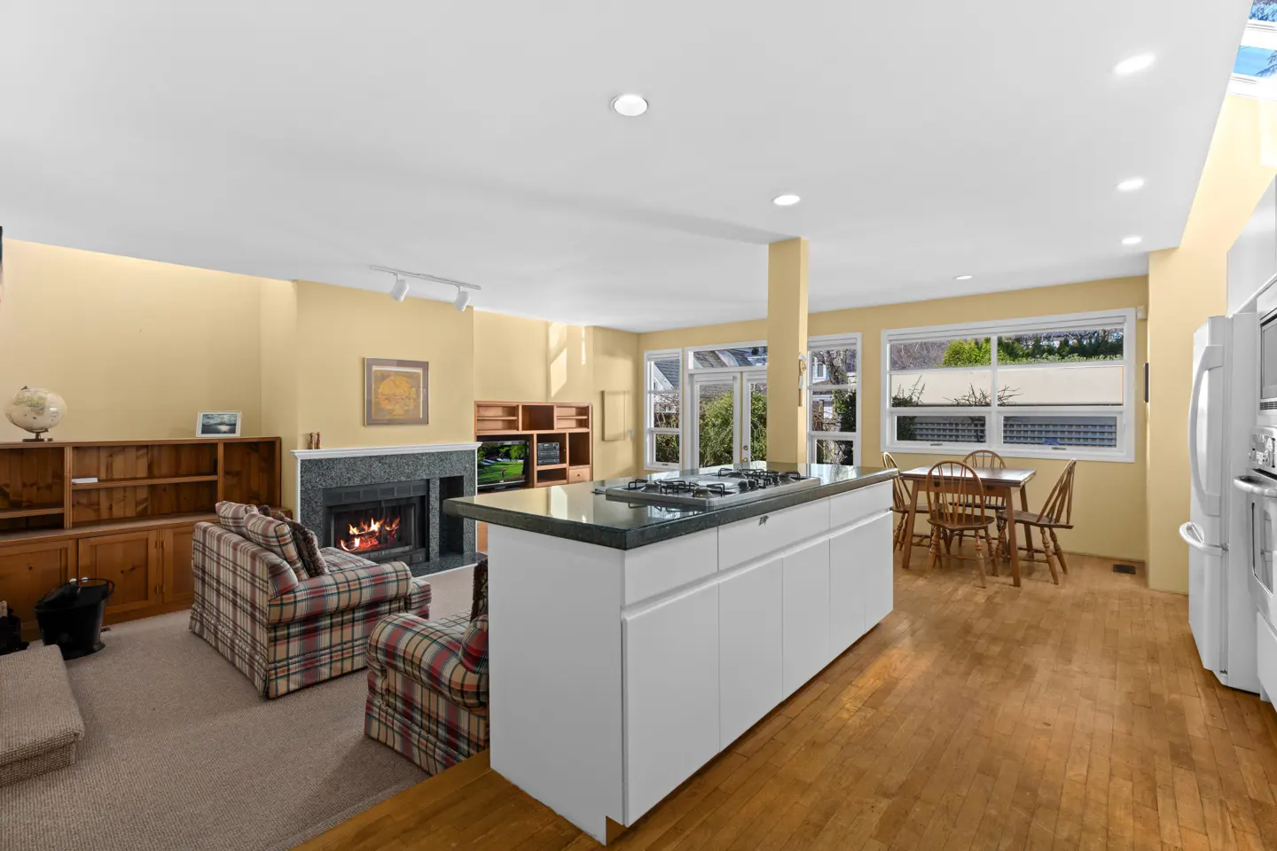Open-concept living space with a kitchen island, fireplace, plaid armchairs, and a dining table near windows.