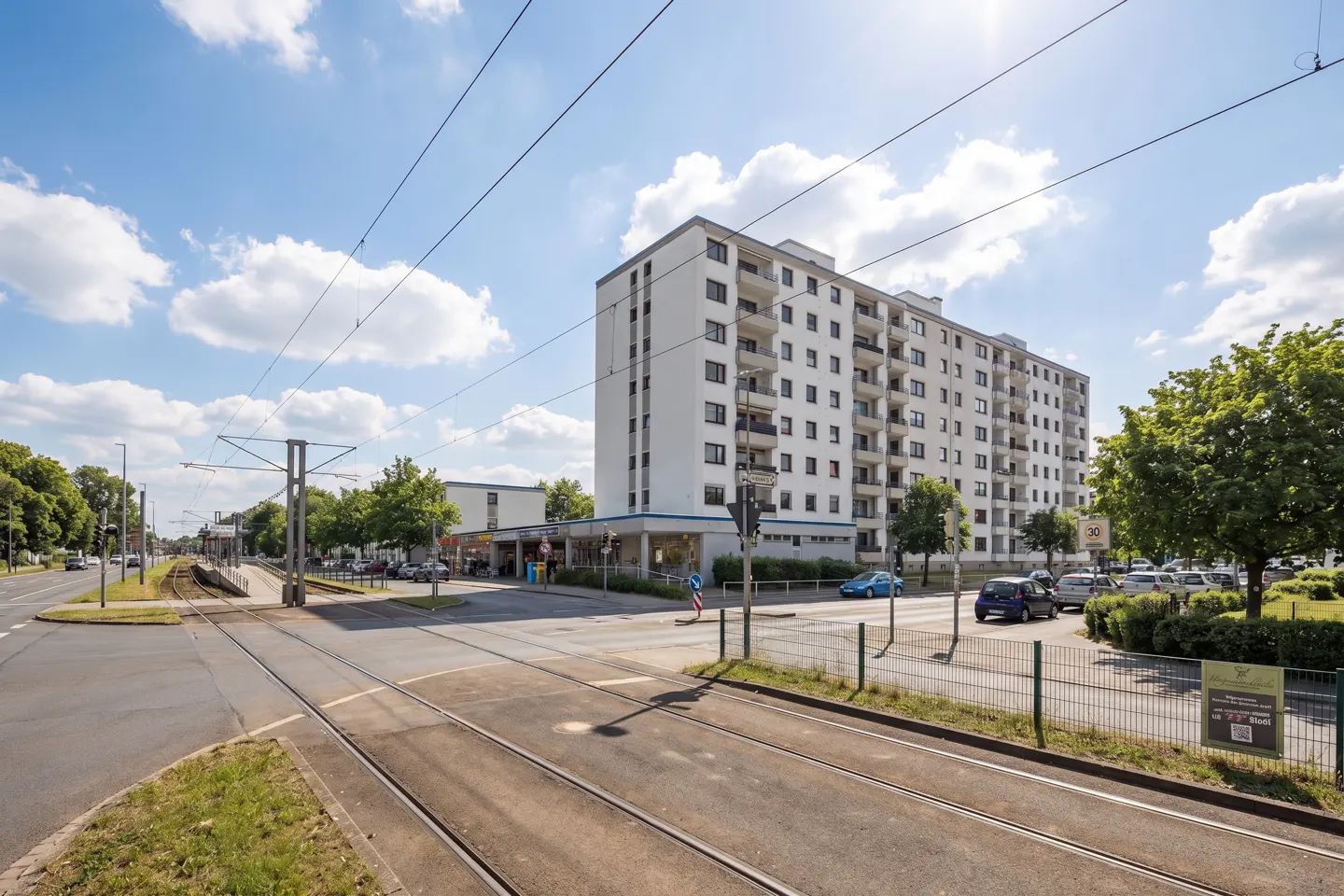 Street view of a white apartment building with balconies, a tram line crossing the road, and a blue sky with clouds.