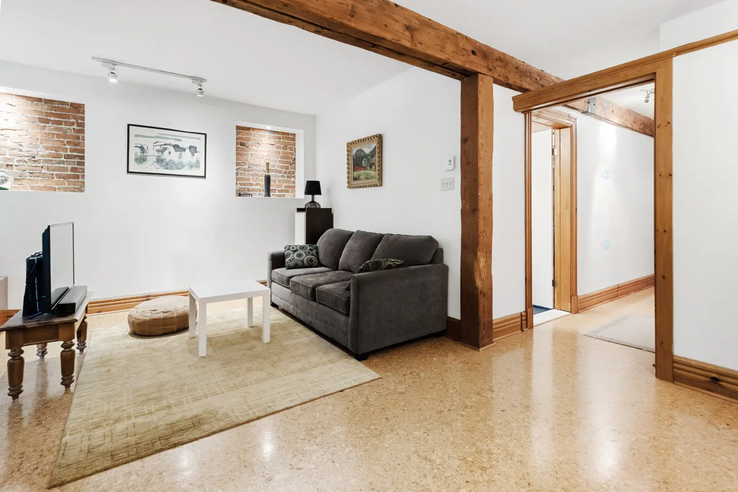 Living room with white walls, exposed brick, and wooden beams. A gray sofa sits on a beige rug, with a white coffee table. A doorway leads to another room.