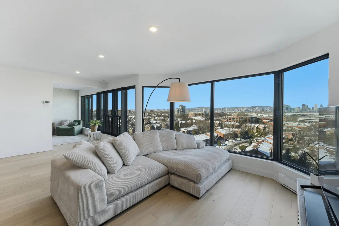 Bright living room with a gray sectional sofa, floor lamp, and large windows overlooking a city skyline.