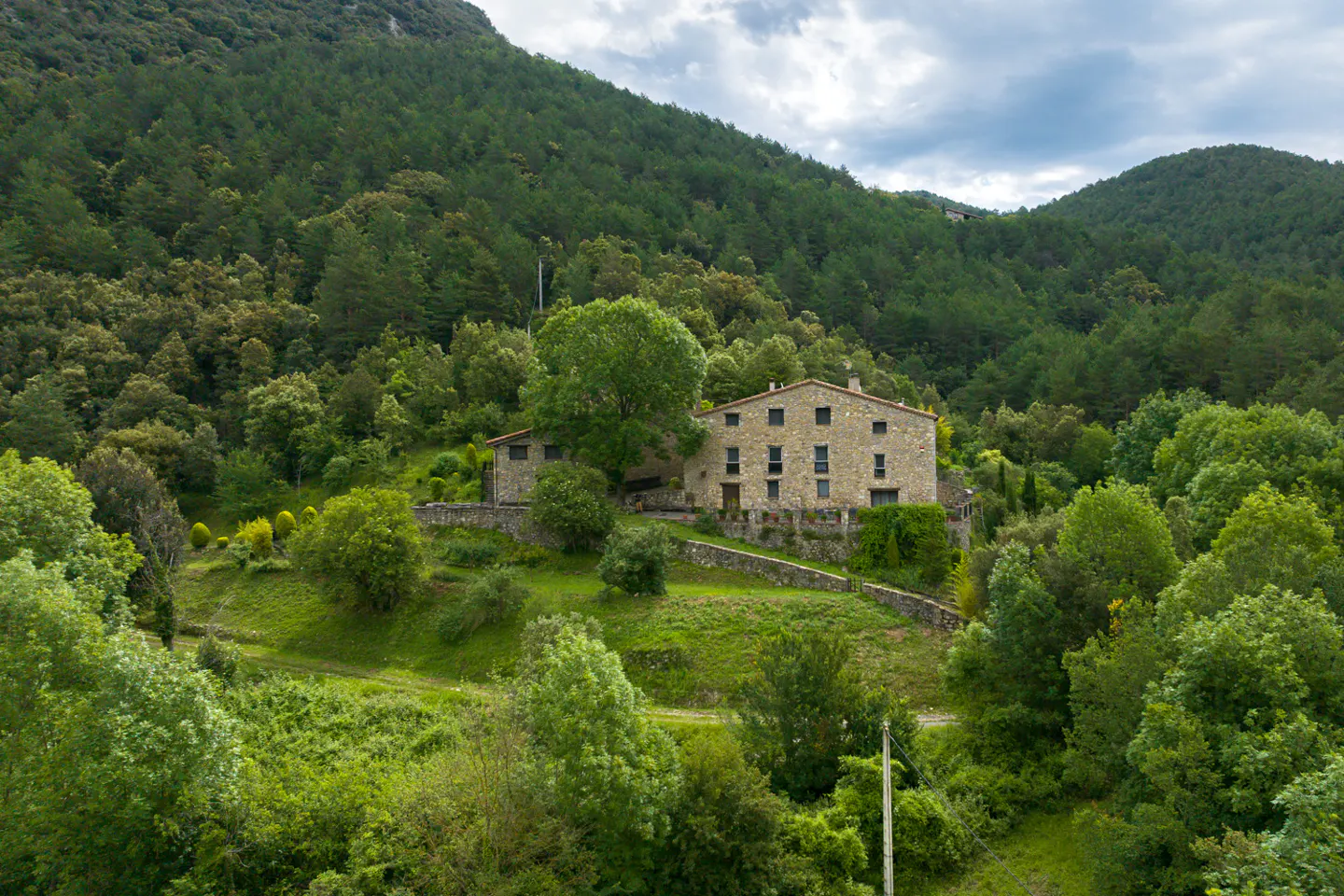 Stone house nestled on a green hillside, surrounded by lush trees and a mountain backdrop.