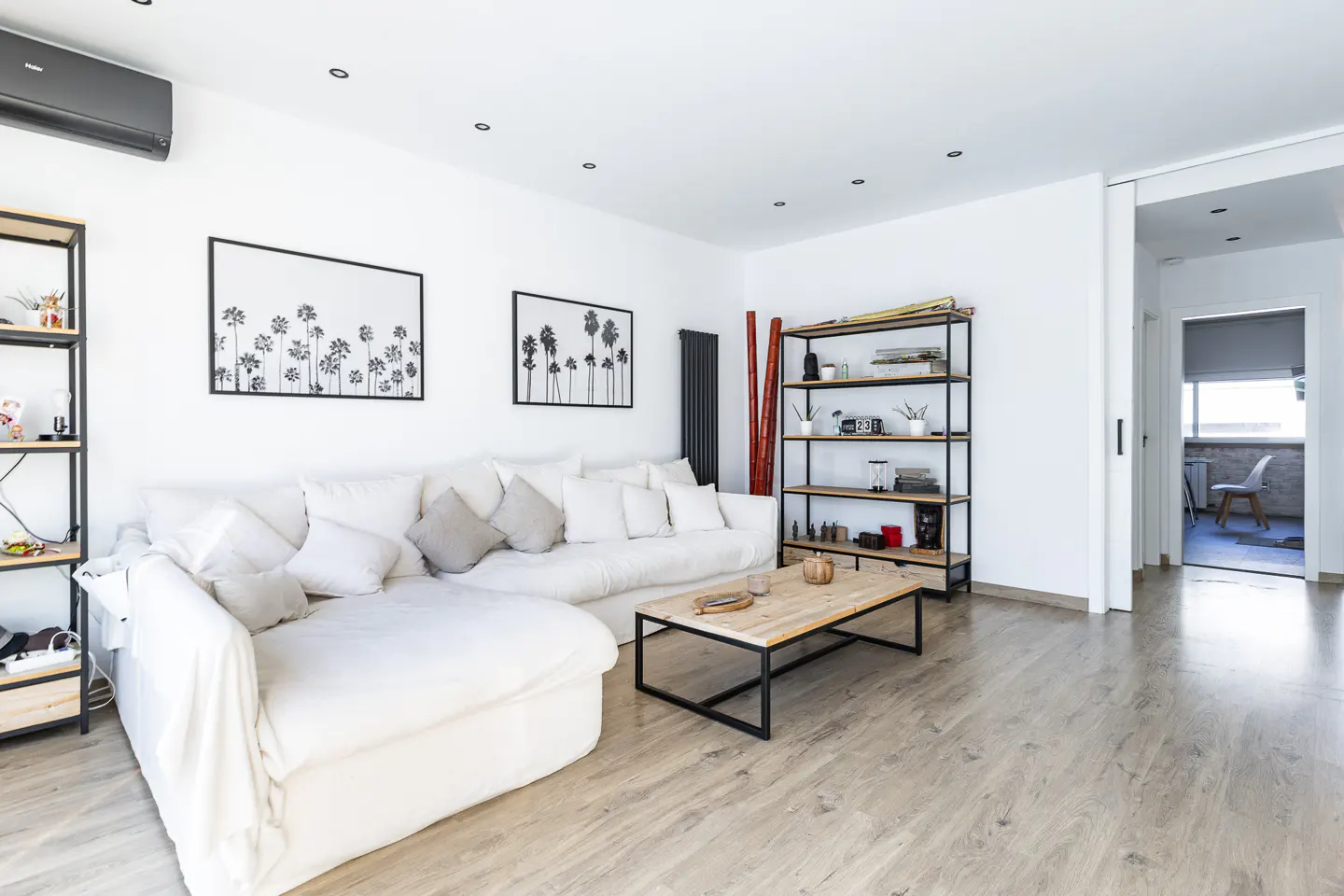 Bright living room with white walls, light wood floors, and a large white sofa. Black framed palm tree art hangs above the sofa. A wood and metal coffee table sits in front.