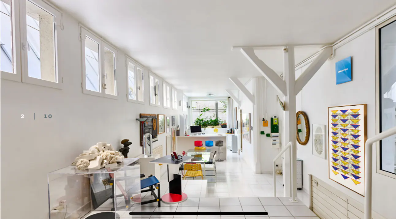 Bright, white loft interior with art. Windows line the left wall, leading to a kitchen area with colorful stools. Stairs on the right.