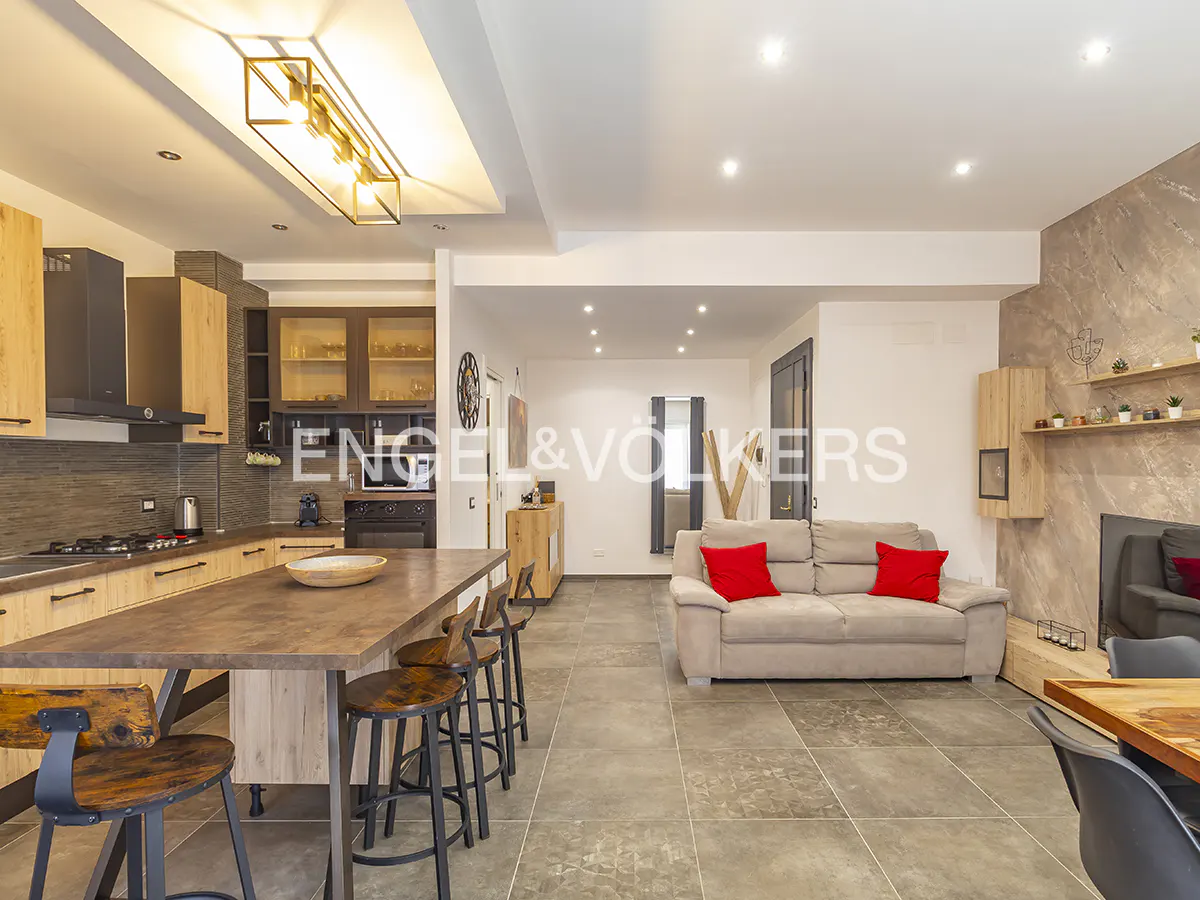 Modern open-plan kitchen and living area with a wooden island, bar stools, beige sofa with red pillows, and tiled floors.