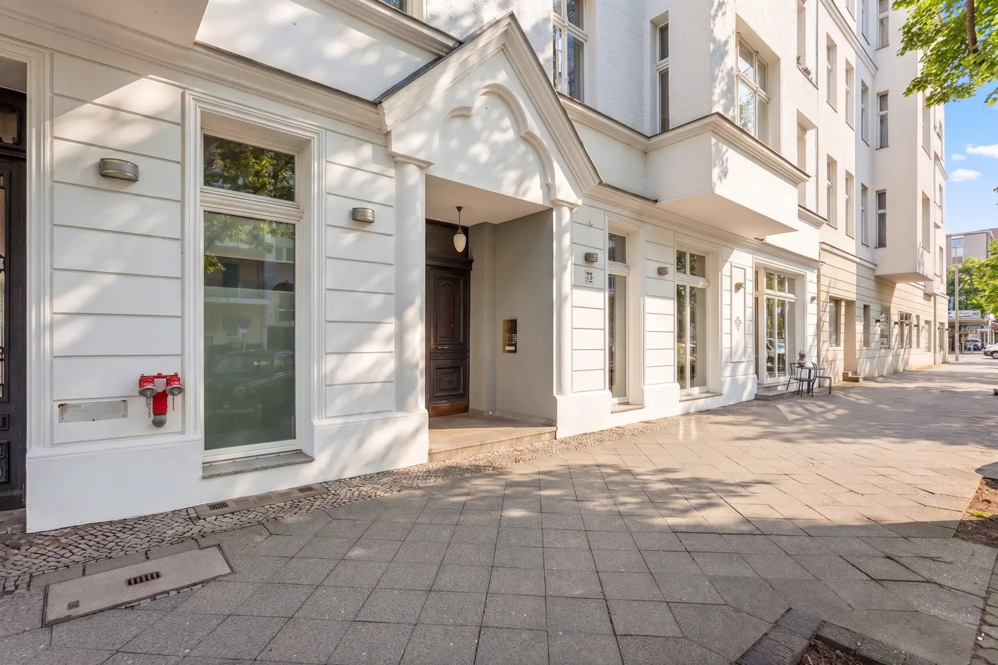 Exterior of a white building with a dark wood door and large windows on a gray tiled sidewalk.