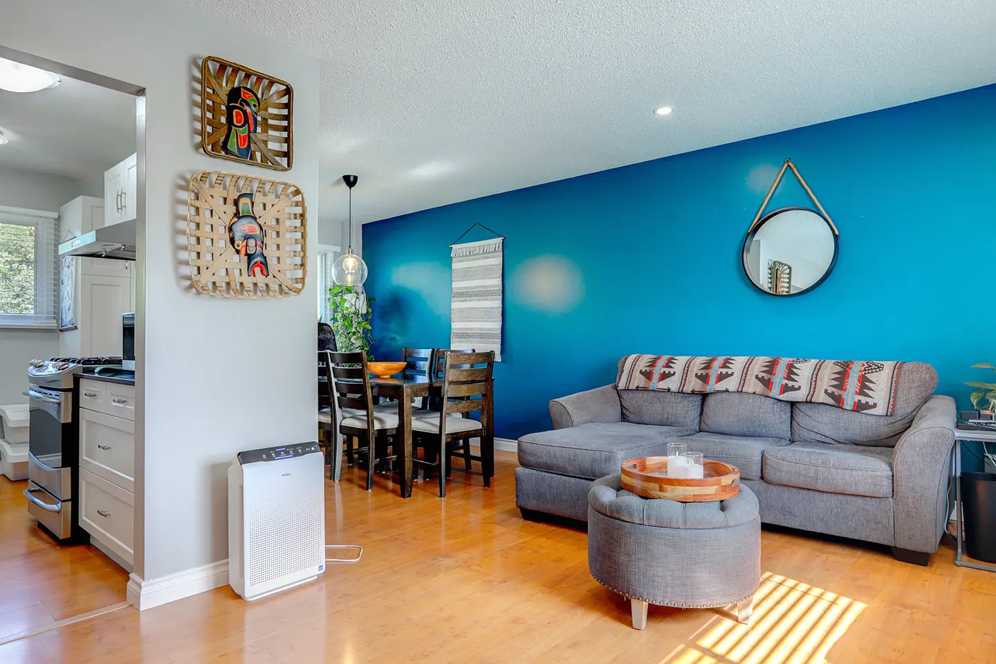 Living room with blue wall, gray sofa, ottoman, and dining table. Woven art hangs on the wall. Hardwood floors and natural light.