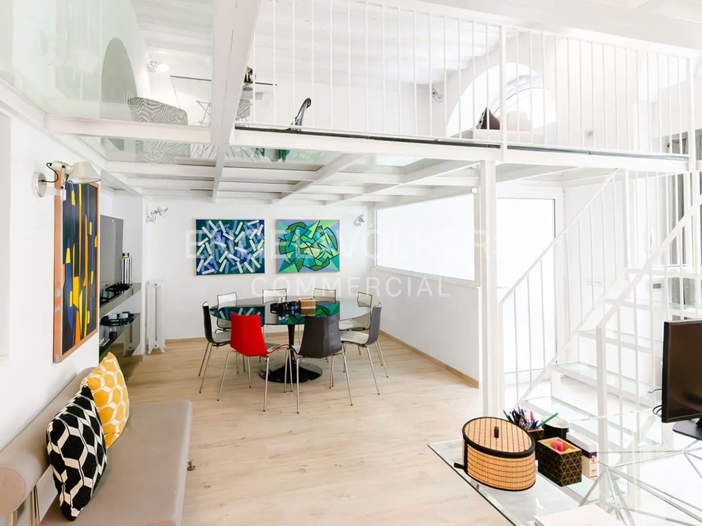 Bright, modern loft apartment with white walls, wood floors, and a glass-enclosed mezzanine level. Dining table and colorful art.