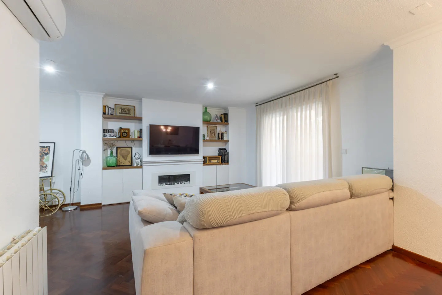 Bright living room with a beige sofa, dark wood floors, and white built-in shelves with a TV and fireplace. Curtains cover a window.