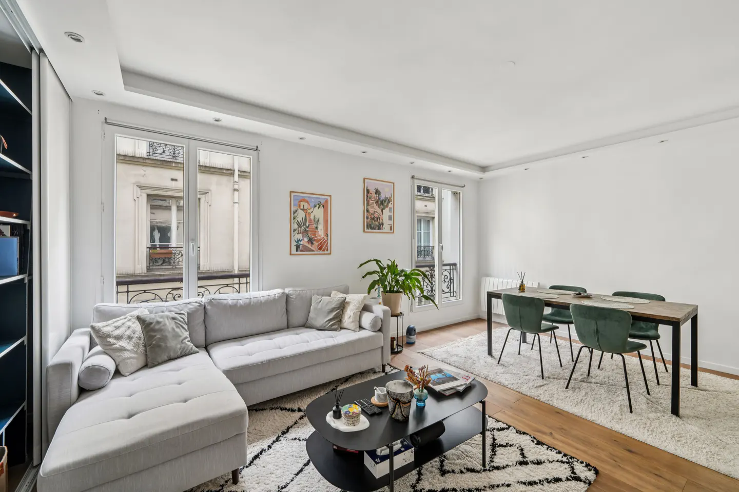 Bright living room with a gray sectional sofa, black coffee table, dining table with green chairs, and large windows.