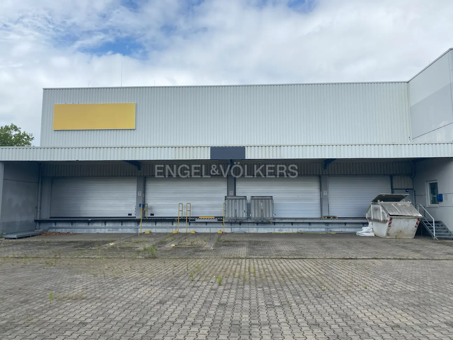 Exterior of a white warehouse with loading docks and an Engel & Völkers sign. A yellow rectangle is above the sign.