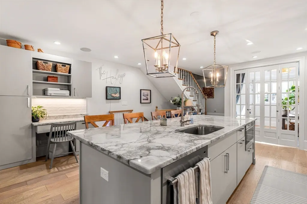 A modern kitchen with a marble island, gray cabinets, and wooden floors. Two pendant lights hang above the island. A staircase is visible in the background.